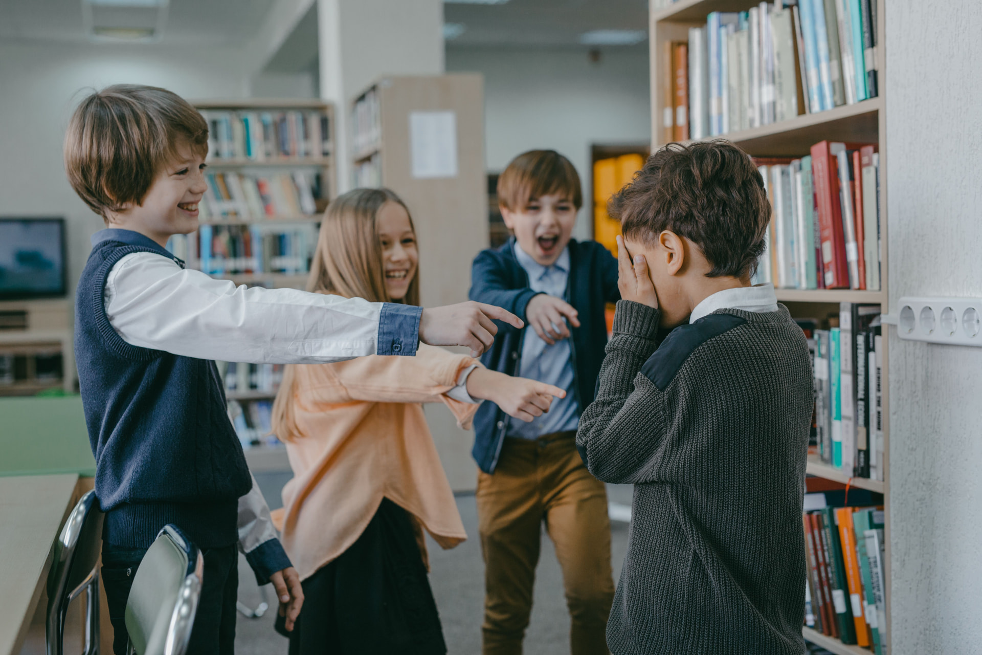 A schoolboy covering his face while other children point and laugh in a school library