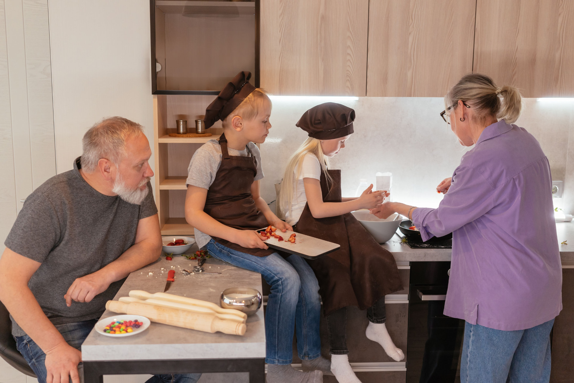 Grandparents baking in a kitchen with two young grandchildren wearing chef hats